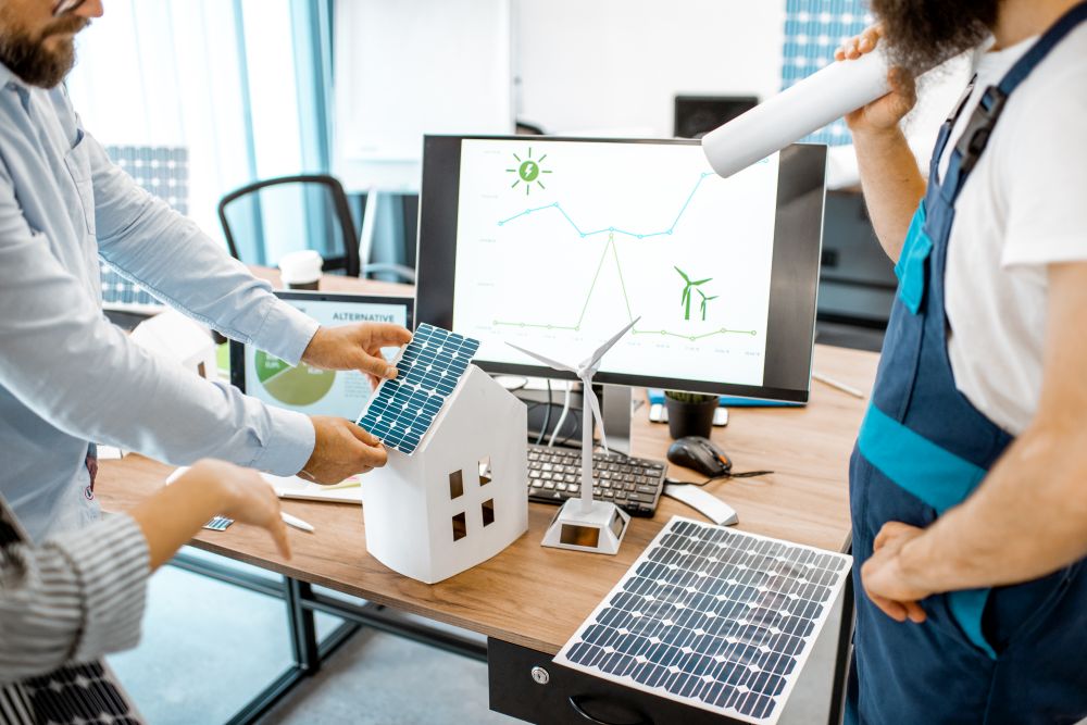 Three people discuss smart energy management, focusing on solar panels and wind turbines at a desk with model houses, charts on a computer screen, and renewable energy equipment.