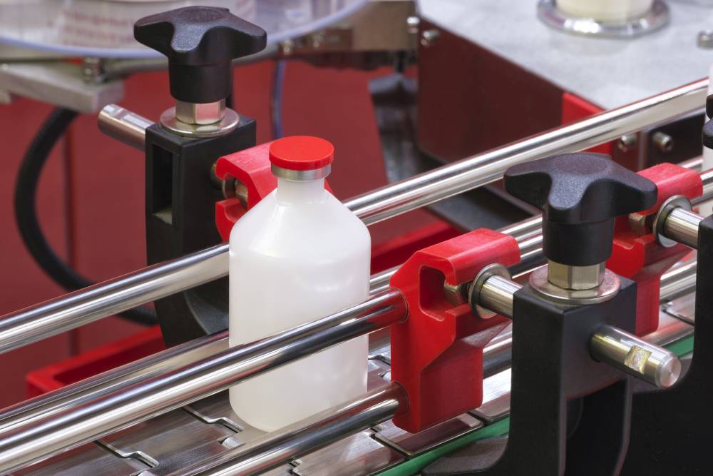 A white plastic bottle with a red cap is held by clamps on a conveyor system in an industrial manufacturing or packaging facility.